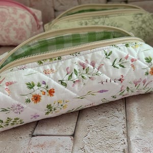 May include: A quilted fabric pencil case featuring a white base with a colourful wildflower print. The pouch has a beige zip closure and an interior lining with a green and white gingham check pattern. The exterior is stitched in a diamond pattern.