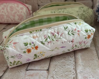 Puede incluir: Un estuche de tela acolchada con base blanca y un estampado de flores silvestres de colores. El estuche tiene una cremallera beige y un forro interior con un patrón de cuadros vichy verdes y blancos. El exterior presenta un diseño de rombos.