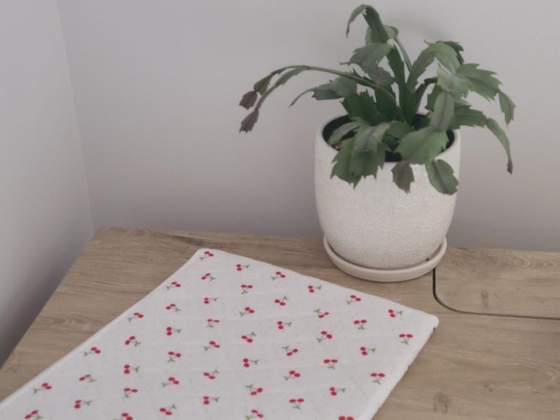 A potted plant with green leaves sits on a wooden surface, accompanied by a white cloth with red cherries on it.