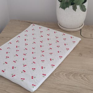 a wooden table with a white cloth featuring a repeating pattern of red cherries, placed next to a white ceramic pot with a plant inside.