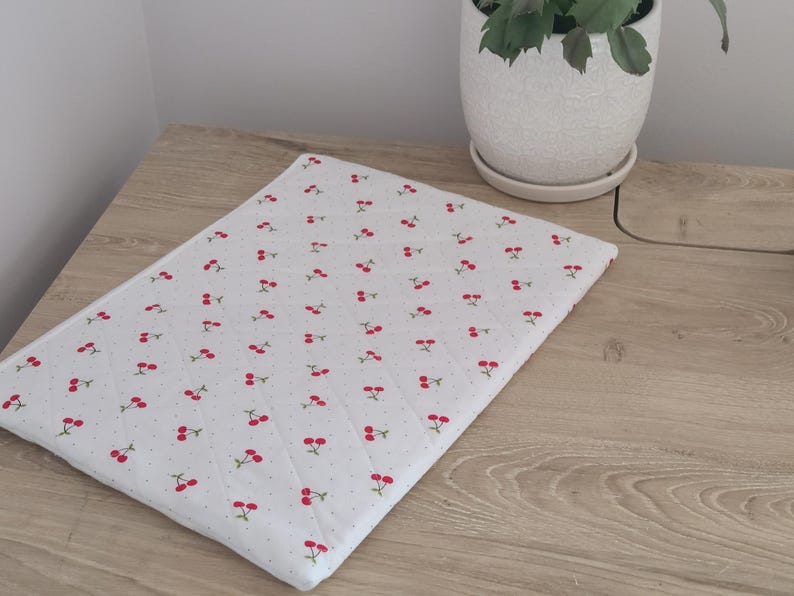 a wooden table with a white cloth featuring a repeating pattern of red cherries, placed next to a white ceramic pot with a plant inside.
