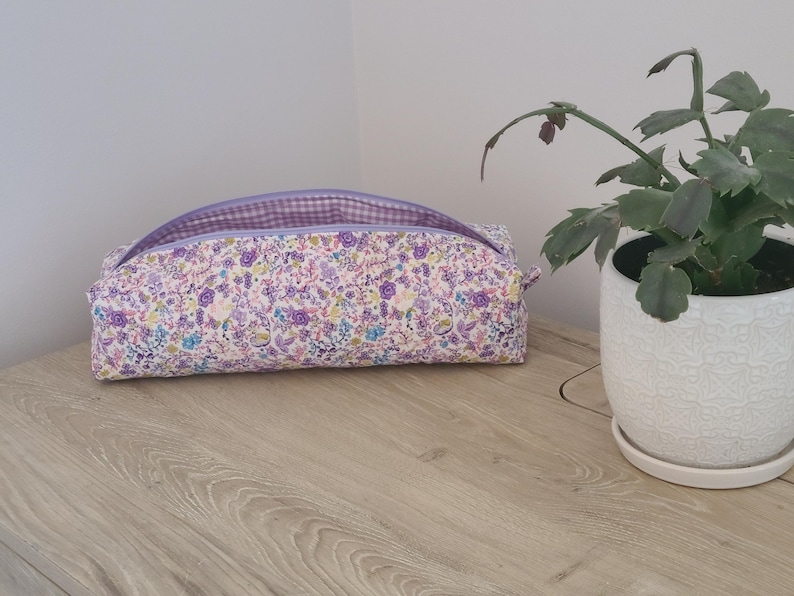 A floral patterned pencil case sits on a wooden surface next to a potted plant.
