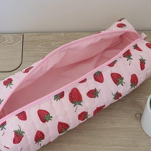 May include: A pink, quilted pencil case with a strawberry pattern. The case is open, revealing a pink interior. The exterior features red strawberries with green leaves and white polka dots. A pink zipper runs along the top.