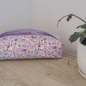A floral patterned bag with a purple zipper sits on a wooden surface next to a potted plant.