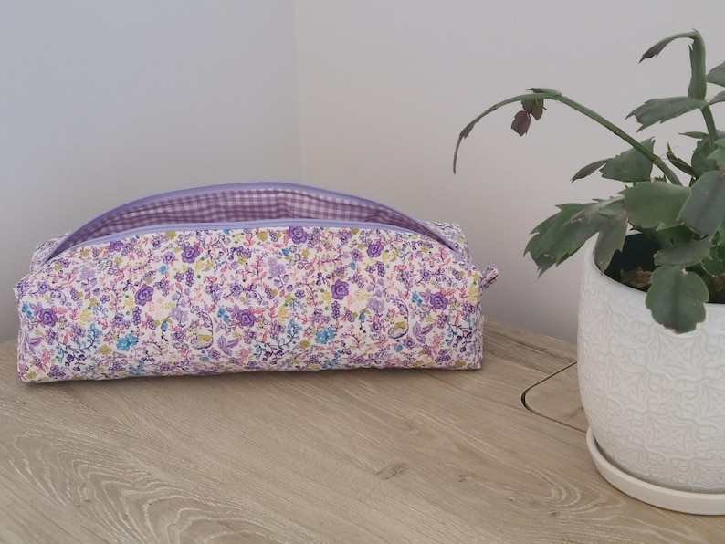 A floral patterned bag with a purple zipper sits on a wooden surface next to a potted plant.