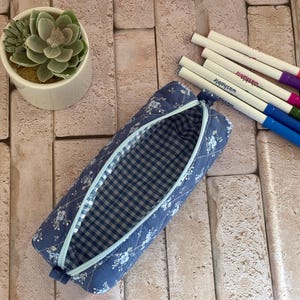 May include: A blue fabric pencil case with a white floral pattern and a light blue zipper. The interior is lined with a blue and white gingham check fabric. Next to the case are four felt-tip markers in pink, purple, green, and blue.