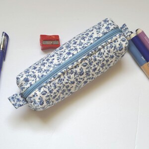 A blue and white floral pencil case, a blue pen, a red pencil sharpener, and a set of colored pencils are arranged on a white surface.