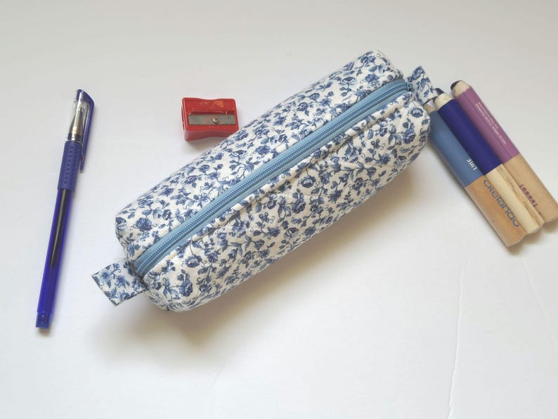 A blue and white floral pencil case, a blue pen, a red pencil sharpener, and a set of colored pencils are arranged on a white surface.