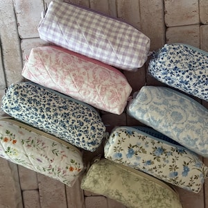Puede incluir: Una colecci&oacute;n de ocho estuches rectangulares de tela acolchada con varios patrones, incluyendo cuadros vichy lavanda, toile floral rosa, estampados florales en azul y blanco, y un estuche crema con peque&ntilde;as flores silvestres.
