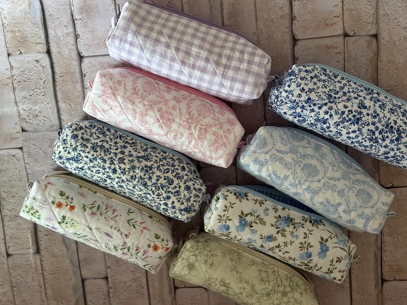 Puede incluir: Una colecci&oacute;n de ocho estuches rectangulares de tela acolchada con varios patrones, incluyendo cuadros vichy lavanda, toile floral rosa, estampados florales en azul y blanco, y un estuche crema con peque&ntilde;as flores silvestres.