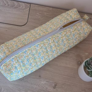 May include: A rectangular pencil case with a floral pattern in yellow, blue, and white. The case has a silver zipper running along the top and is lying on a wooden surface. A small potted succulent is next to the case.