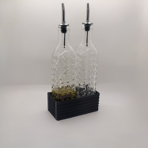 以下が含まれることがあります： 銀色の注ぎ口と黒のテクスチャ加工されたベースが付いた透明なガラス瓶2本。瓶は薄い黄色の液体で満たされています。