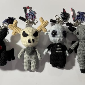 May include: A collection of handmade crochet keychains featuring various spooky characters. The keychains include a white figure with a red tie, a black bat-like creature, a deer-skull figure, a skull figure, a gray figure, and a character in an orange jumpsuit.