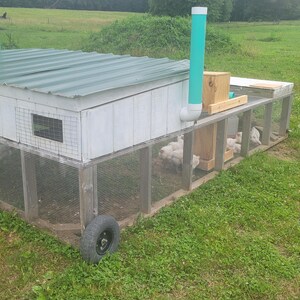 May include: A white and green chicken coop with a metal roof, wire mesh sides, and two teal ventilation pipes. The coop is on wheels and sits in a grassy field. Several chickens are visible inside.