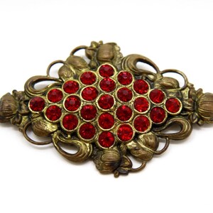 May include: An antique-style brooch with a central cluster of red rhinestones. The brooch has an ornate, filigree design in a bronze tone, with floral and scroll details. The overall shape is elongated and symmetrical, suitable for adding a vintage touch to any outfit.