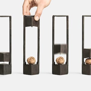 Può includere: Un schiaccianoci in metallo nero con una noce all'interno. Lo schiaccianoci è mostrato in quattro fasi: aperto, con una noce all'interno, con la noce che viene schiacciata e con la noce schiacciata all'interno.