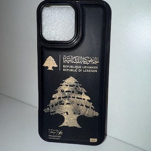 May include: Black phone case with a gold emblem of a cedar tree and text in Arabic and English. The case has a camera cutout and a smooth, textured finish. The text reads "REPUBLIQUE LIBANAISE REPUBLIC OF LEBANON" and "PASSEPORT PASSPORT".