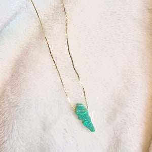 Puede incluir: Un collar de cadena dorada con un colgante verde claro. El colgante, de forma irregular, est&aacute; adornado con purpurina iridiscente. El collar se muestra sobre un fondo blanco texturizado.