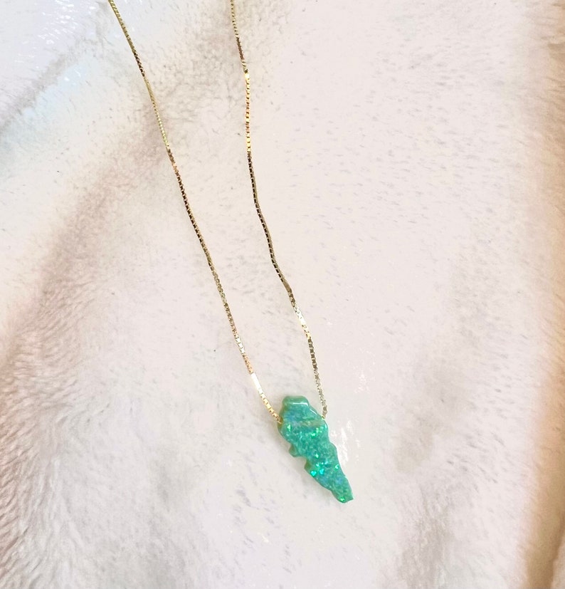 Puede incluir: Un collar de cadena dorada con un colgante verde claro. El colgante, de forma irregular, est&aacute; adornado con purpurina iridiscente. El collar se muestra sobre un fondo blanco texturizado.
