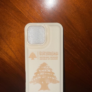 May include: Off-white phone case featuring a beige cedar tree design and text in Arabic and English. The case has a camera cutout and a passport stamp graphic at the bottom. The case is made of a smooth material.