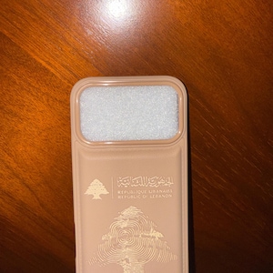 May include: A tan phone case featuring the text "REPUBLIQUE LIBANAISE REPUBLIC OF LEBANON" in English and Arabic, along with a cedar tree emblem. The case has a rectangular opening at the top and a textured surface.