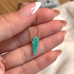 Puede incluir: Un collar dorado con un colgante con forma de pa&iacute;s, con una piedra azul-verde brillante e iridiscente. El colgante cuelga de una delicada cadena. El collar se sostiene en una mano.