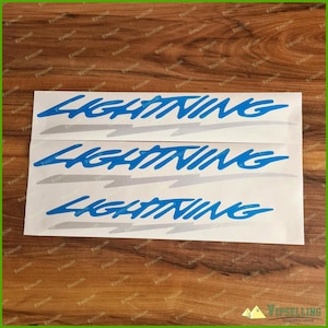 May include: Three blue "LIGHTNING" decals with a stylized font and a lightning bolt design. The decals are on a white background, with a grey shadow effect. The background is a wooden surface.