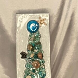 May include: A white canvas with a blue and green mosaic Christmas tree made of seashells and glass. The tree is topped with a blue ornament and a gold starfish.