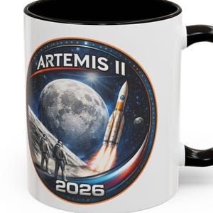 以下が含まれることがあります： 黒い内側とハンドルが付いた白いセラミックマグ。マグには「ARTEMIS II」の文字と「2026」の年号が入った円形のデザインが施されています。デザインには、月に向かって打ち上げられるロケットと、月の表面にいる宇宙飛行士が含まれています。