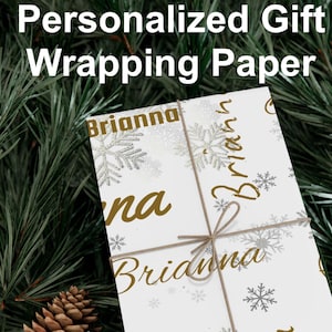 May include: White gift wrapping paper with the name "Brianna" in gold script and snowflake accents. The paper is tied with twine and set against a backdrop of pine needles and pine cones. Text at the top reads "Personalized Gift Wrapping Paper."
