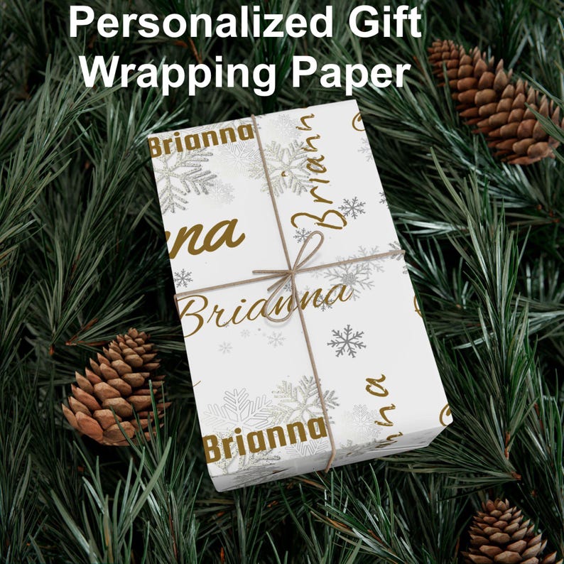 May include: White gift wrapping paper with the name "Brianna" in gold script and snowflake accents. The paper is tied with twine and set against a backdrop of pine needles and pine cones. Text at the top reads "Personalized Gift Wrapping Paper."