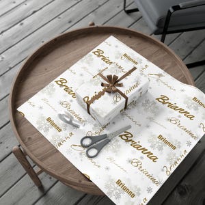 May include: A wrapped gift box with a brown ribbon and the name "Brianna" printed on the white wrapping paper. The paper also features snowflakes and the name "Brianna" in a repeating pattern. Scissors and tape are on the table.