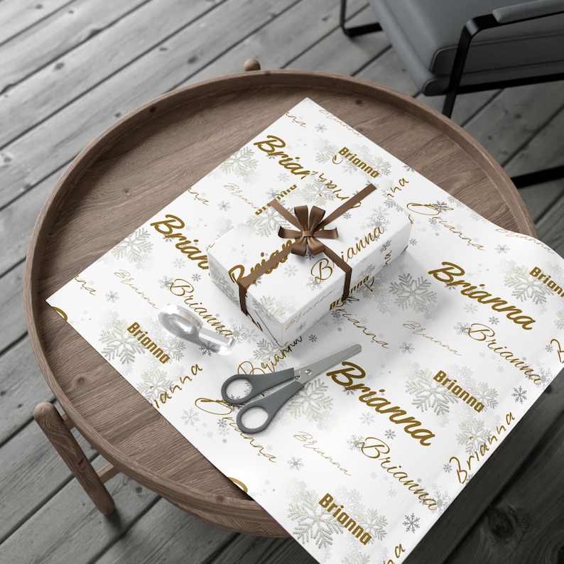 May include: A wrapped gift box with a brown ribbon and the name "Brianna" printed on the white wrapping paper. The paper also features snowflakes and the name "Brianna" in a repeating pattern. Scissors and tape are on the table.