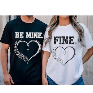 May include: Black and white t-shirts with matching heart designs. The black shirt says "BE MINE." and the white shirt says "FINE." Both shirts feature a large heart with smaller hearts around it.