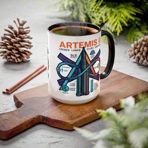 May include: A white ceramic mug with a black handle and rim, featuring a diagram of the Artemis II mission. The design includes the text "ARTEMIS II CREWED LUNAR FLYBY MISSION" and other mission details in teal, orange, and purple.