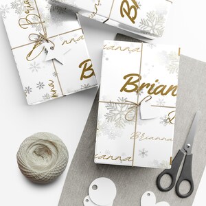 May include: White gift boxes wrapped in paper with gold snowflakes and the names "Brian" and "Brianna" printed on them. The boxes are tied with twine and adorned with star and heart-shaped tags. A ball of twine and scissors are also present.