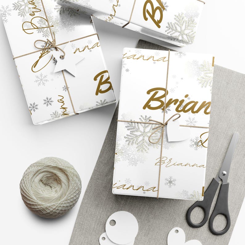 May include: White gift boxes wrapped in paper with gold snowflakes and the names "Brian" and "Brianna" printed on them. The boxes are tied with twine and adorned with star and heart-shaped tags. A ball of twine and scissors are also present.