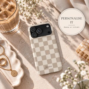 May include: A beige and white checkered phone case featuring a black camera module and the name Jessica printed in a cursive script at the bottom. The case has a smooth finish and is displayed alongside a glass of iced coffee and jewelry.
