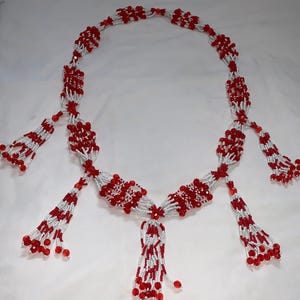 Peut inclure: Un collier de perles rouges et blanches, présenté sur un fond blanc. Le collier présente un motif central et plusieurs glands perlés. Les perles sont petites et rondes, créant un motif détaillé. Le collier est probablement fait à la main.