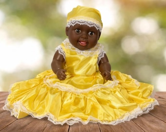 May include: A dark-skinned baby doll wearing a yellow dress and matching head covering, trimmed with white lace. The doll is smiling, and the background is a blurred green and brown.