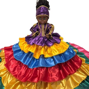 May include: A doll wearing a purple and gold dress with a matching head covering. The doll's skirt is made of layers of colorful satin in red, blue, yellow, and green. The doll has dark skin and black hair.