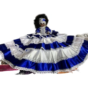 May include: A doll wearing a layered blue and white dress with silver trim and lace edging. The doll has dark skin, black curly hair, and a matching blue and silver headpiece. The dress has multiple tiers of fabric.