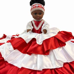 May include: A doll with dark skin wearing a red and white dress. The dress has multiple layers of satin fabric. The doll has black curly hair and a red headband with white trim.