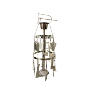 May include: A silver-coloured, hanging wind chime with a decorative design. The chime has multiple tiers of metal shapes suspended by chains, with a central column and a top hanging element. The shapes include various geometric and abstract forms.