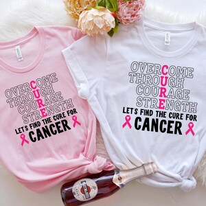 May include: Two pink and white t-shirts with the word "CURE" spelled out vertically with the letters in pink. The text reads "Overcome Through Courage Strength Let's Find the Cure for Cancer" with a pink ribbon on either side of the word "Cancer".