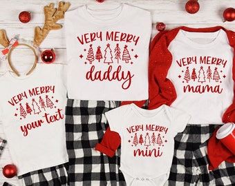 Op de afbeelding: Witte kerstpyjama's met rode tekst die "Very Merry" en "daddy", "mama", "mini" en "your text" zegt. De pyjama's hebben een zwart-wit geruit patroon.
