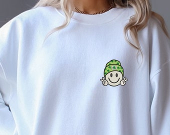 Puede incluir: Sudadera blanca con un pequeño bordado de una carita sonriente con un gorro verde con tréboles. La carita sonriente hace un gesto de paz con las manos. La sudadera es de cuello redondo.