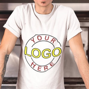 May include: A man wearing a white t-shirt with a red circle and the text "YOUR LOGO HERE" in yellow. He is standing in a kitchen with a cutting board and a tomato.