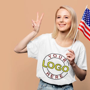 May include: A woman wearing a white t-shirt with a red circle and the text "YOUR LOGO HERE" in yellow. She is holding an American flag in her right hand and giving a peace sign with her left hand.