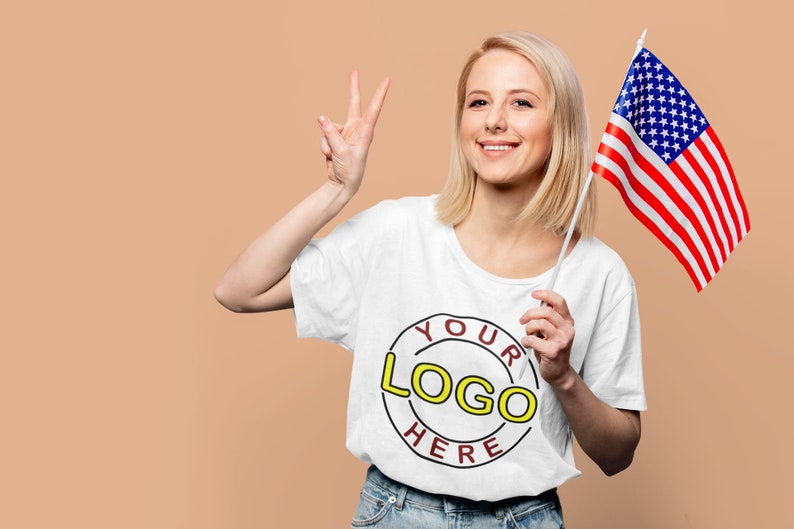May include: A woman wearing a white t-shirt with a red circle and the text "YOUR LOGO HERE" in yellow. She is holding an American flag in her right hand and giving a peace sign with her left hand.
